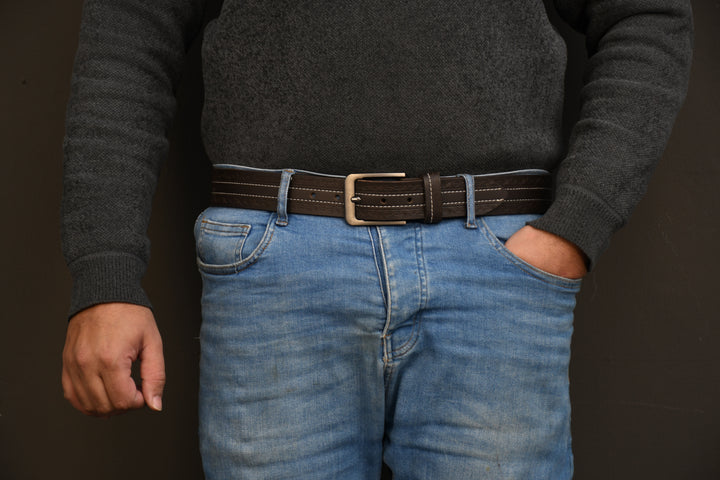 Person wearing a dark gray sweater, blue jeans, and a brown belt with a buckle on a dark background.